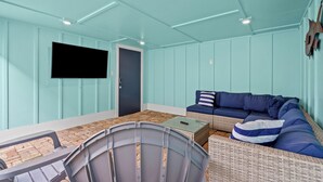 A room with teal walls, a flat-screen TV mounted on the wall, a dark door, a blue sofa with striped pillows, and a wicker chair. - Island Pride Coastal Cottage - 2 King Beds on the East End! (Galveston)