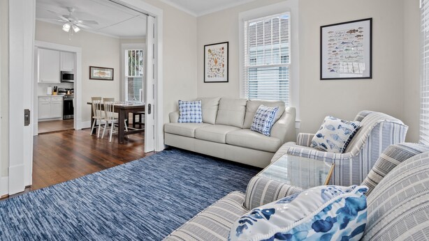 A spacious living room with a beige sofa, a striped armchair, and a blue rug. There is a dining area with a table and chairs, and a kitchen in the background. - Island Pride Coastal Cottage - 2 King Beds on the East End! (Galveston)