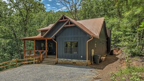 A cabin with a porch, a stone foundation, and a metal roof, surrounded by dense forest. - Holiday House - Pet Friendly | Hot Tub | Modern Mountain Luxury (Blue Ridge)