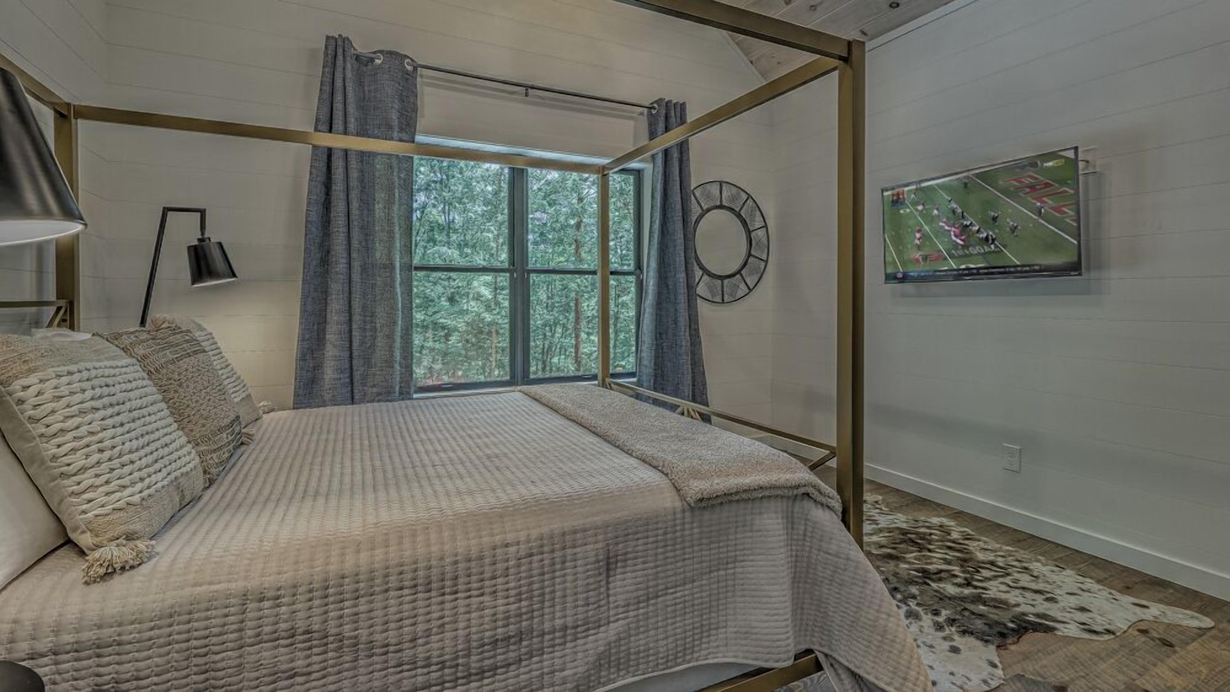 A bedroom with a four-poster bed, a large window with curtains, a round wall clock, and a mounted TV displaying a sports game.