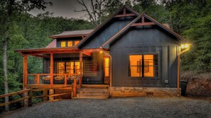 A rustic cabin with a wooden porch, a stone foundation, and a large window with a view of the surrounding forest. - Holiday House - Pet Friendly | Hot Tub | Modern Mountain Luxury (Blue Ridge)