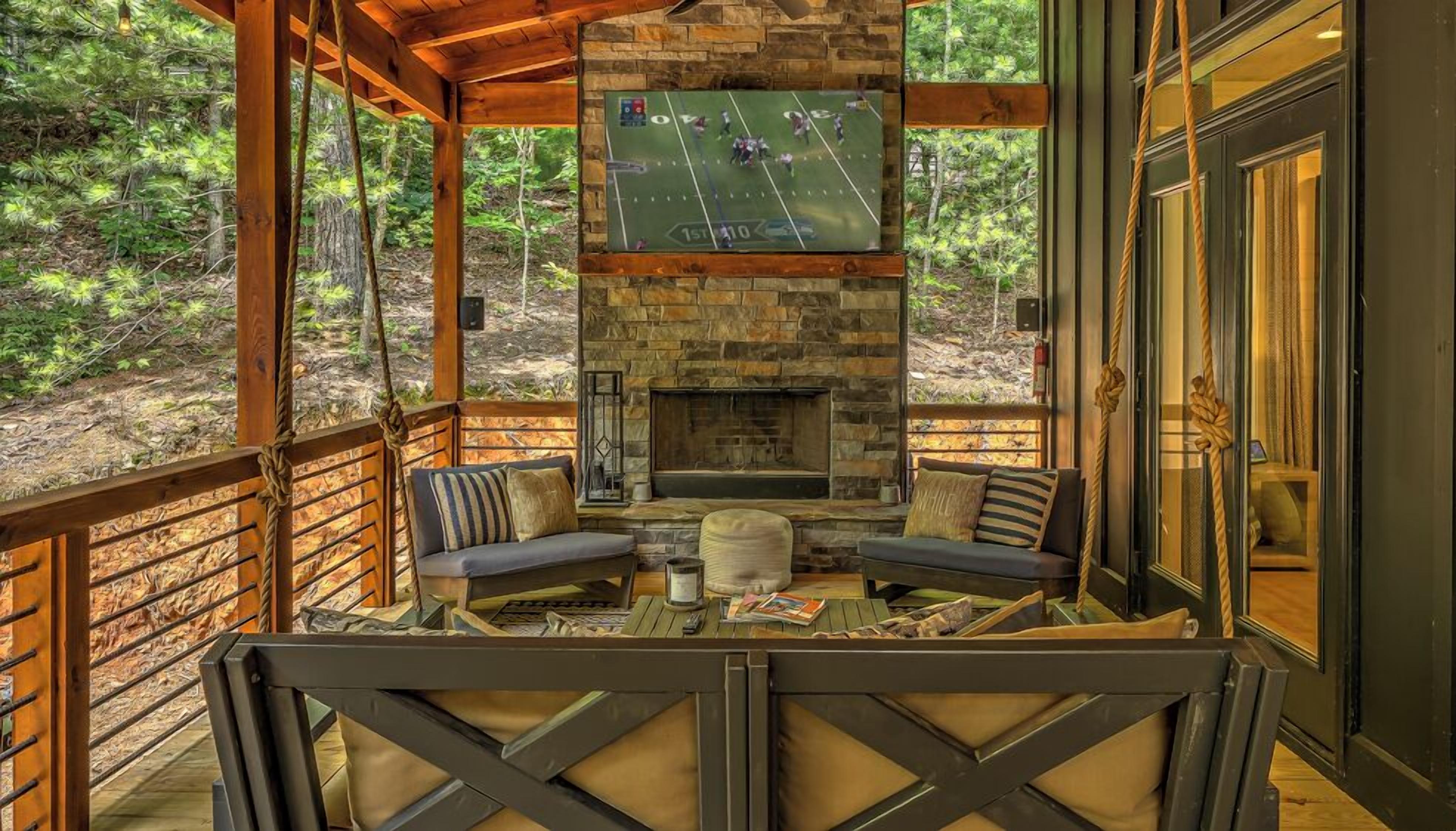 A covered outdoor seating area with a stone fireplace, two sofas, and a television screen displaying a football game.