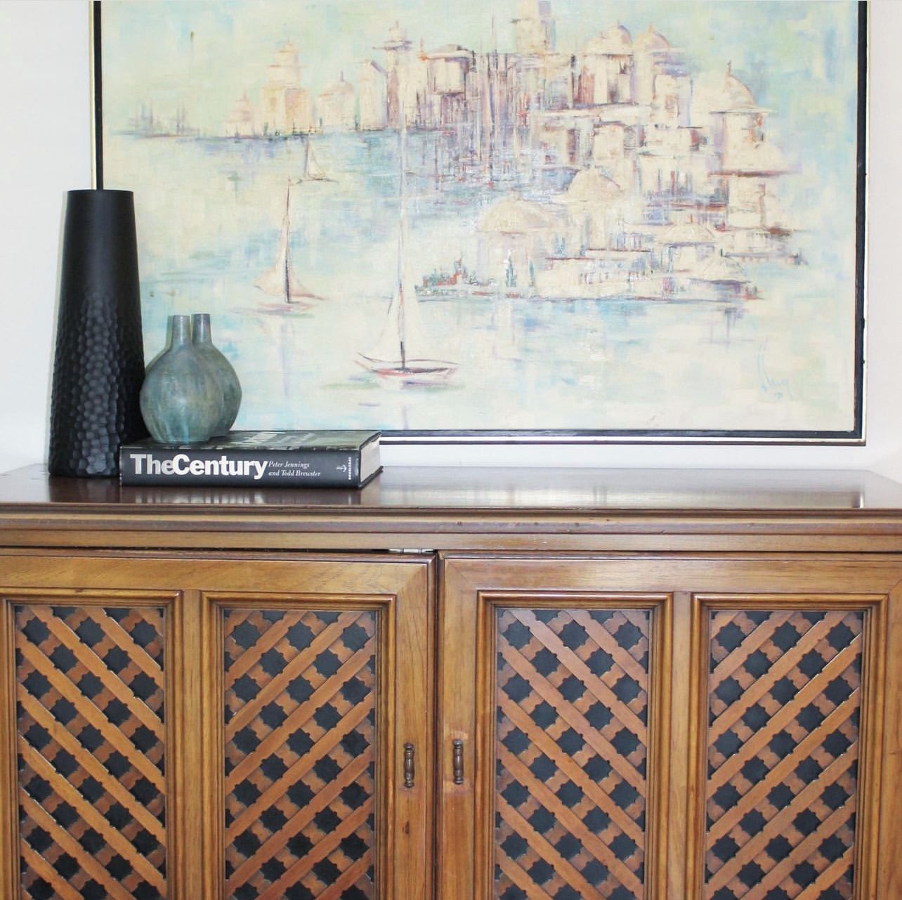 Een lijst met een schilderij van een stadsbeeld met boten op het water, een boek getiteld 'The Century' van Peter Jennings en Todd Drexler, en een houten kastje met een ruitwerk motief.
