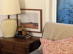 A room with a wooden nightstand, a lamp, a framed picture, a patterned sofa, and a window with a bamboo view.