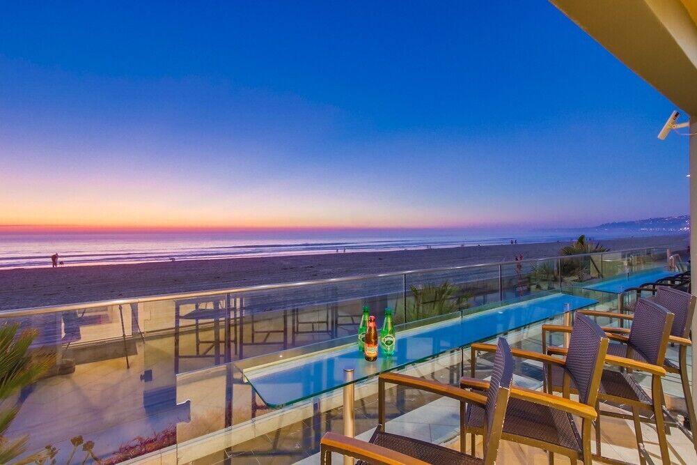 Una piscina infinita con paredes de vidrio y vista a la playa al atardecer.