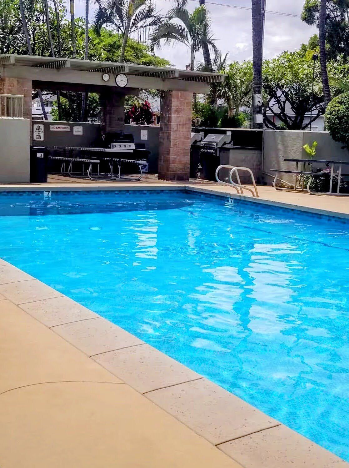 Una piscina con un cartel de prohibido fumar, una parrilla al aire libre y palmeras al fondo.