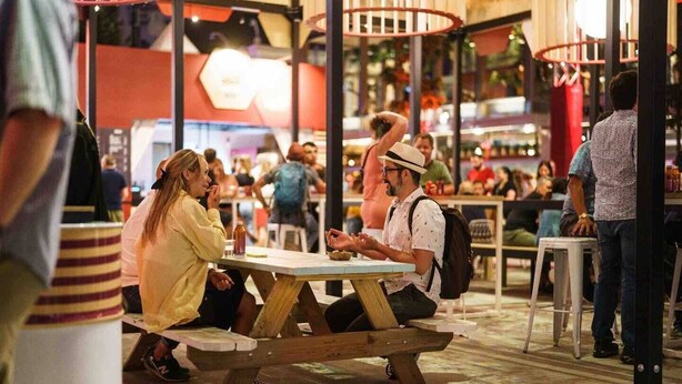 Un espacio al aire libre concurrido para comer, con mesas de picnic de madera y gente sentada y de pie.