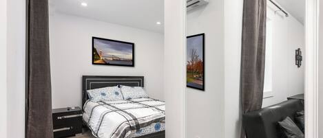 A bedroom with a bed, a nightstand, a dresser, a window with curtains, and an air conditioning unit.