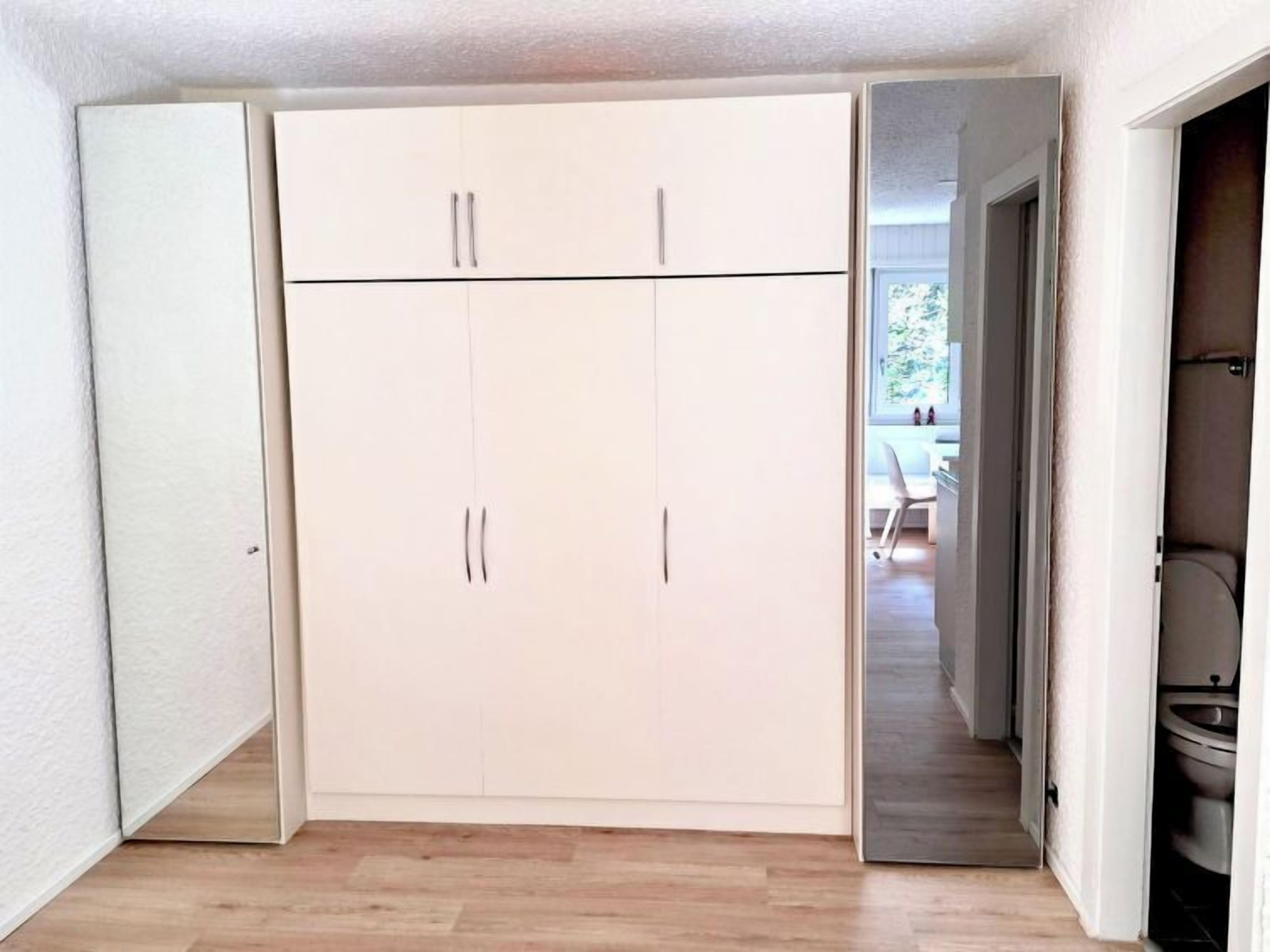 Une pièce dotée d’une grande armoire, d’un plancher en bois et donnant sur un coin repas par une porte ouverte.
