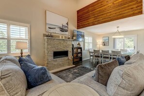 A living room with a stone fireplace, a dining area with a glass table, and a high ceiling with exposed wood beams.