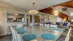 A spacious dining area with a glass table and white chairs, a kitchen in the background, and a loft bedroom with a wooden beam ceiling. - Saltwater Charm | 995 ft. to the Beach | Private Pool (Virginia Beach)