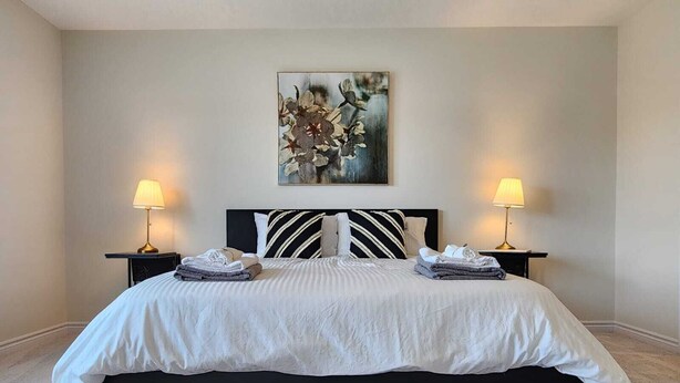 A neatly made bed with a black headboard and white bedding, flanked by two bedside tables with lamps, in a room with a neutral color palette and a single piece of abstract art on the wall. - Cozy Niagara Retreat ,15 min drive to Niagara Falls, 4 bdrms, sleeps up to 9 (Thorold)