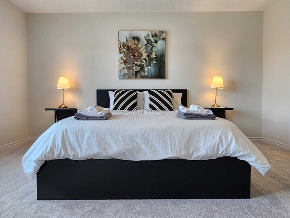 A neatly made bed with a black headboard and white bedding, flanked by two bedside tables with lamps, in a room with a neutral color palette and a single piece of abstract art on the wall.