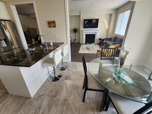 A modern kitchen with a black marble countertop, white bar stools, and a dining area with a glass table and chairs.