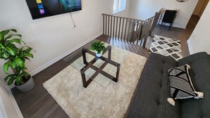 A living room with a TV displaying a profile selection screen, a staircase, a potted plant, a coffee table, and a sofa with a patterned pillow. - Cozy Niagara Retreat ,15 min drive to Niagara Falls, 4 bdrms, sleeps up to 9 (Thorold)