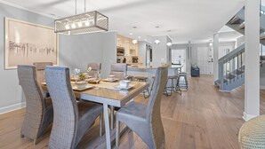 Een ruime eethoek met een houten tafel en geweven stoelen, een keuken op de achtergrond en een trap die naar een hoger niveau leidt.
