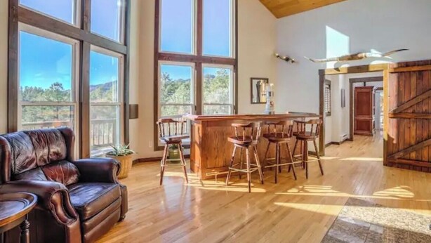 A spacious living area with a large window, a wooden ceiling, and a bar area with stools. - 4BR 4BA multi-family with multiple outdoor & indoor living spaces, hot tub, BBQ. (Colorado Springs)