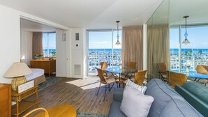 A spacious living area with a large window overlooking a marina, a dining table with chairs, a sofa, and a small desk with a lamp. - Ilikai Marina - Great Location in Waikiki! Stunning Sunset View, Fireworks on Friday! (Honolulu)