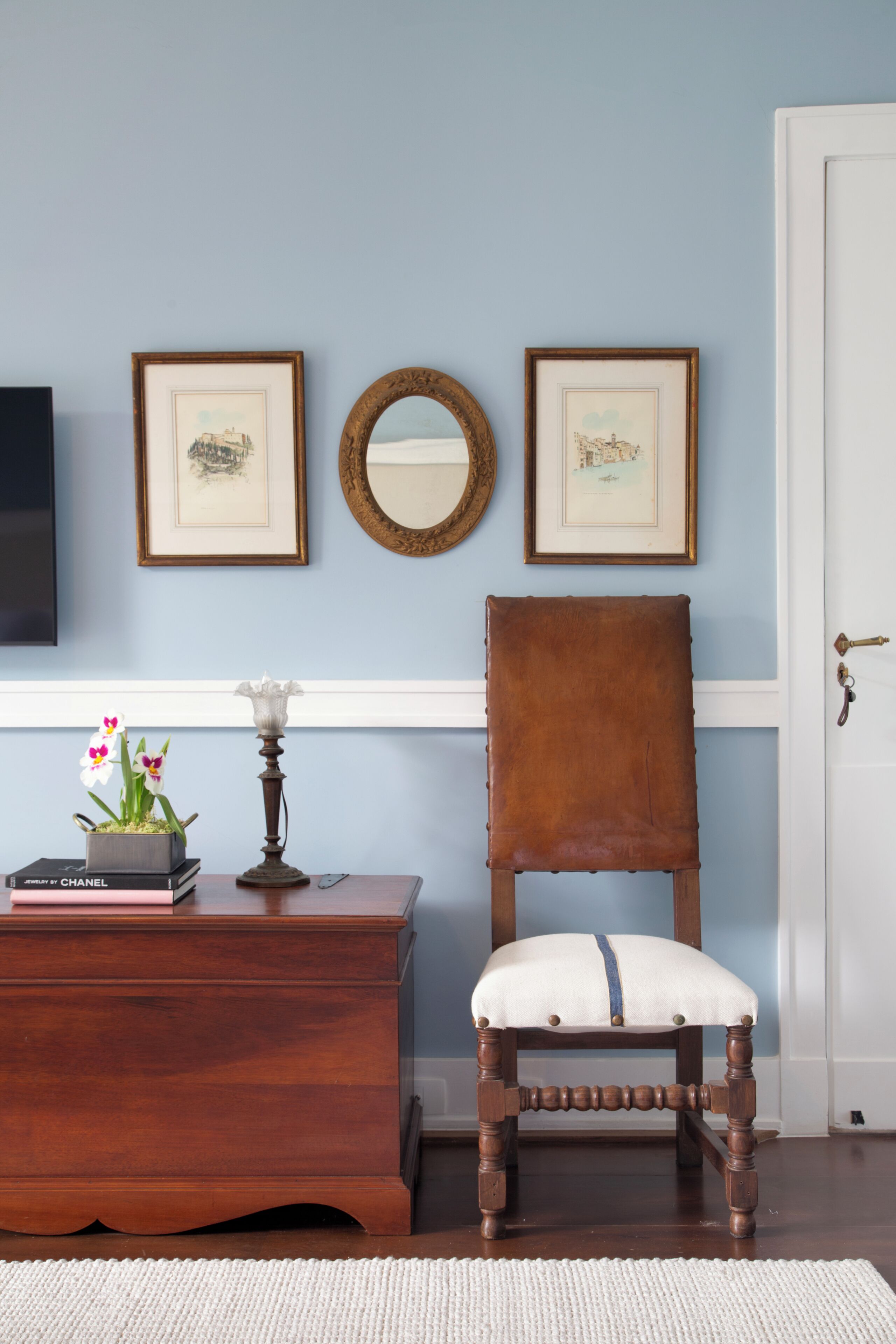 A room with a wooden chair, a small table with books and flowers, framed pictures on the wall, and a mirror.