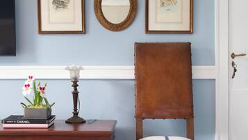 A room with a wooden chair, a small table with books and flowers, framed pictures on the wall, and a mirror.