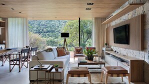 Uma sala de estar moderna com parede de pedra, uma televisão grande e uma área de jantar com vista para a floresta.