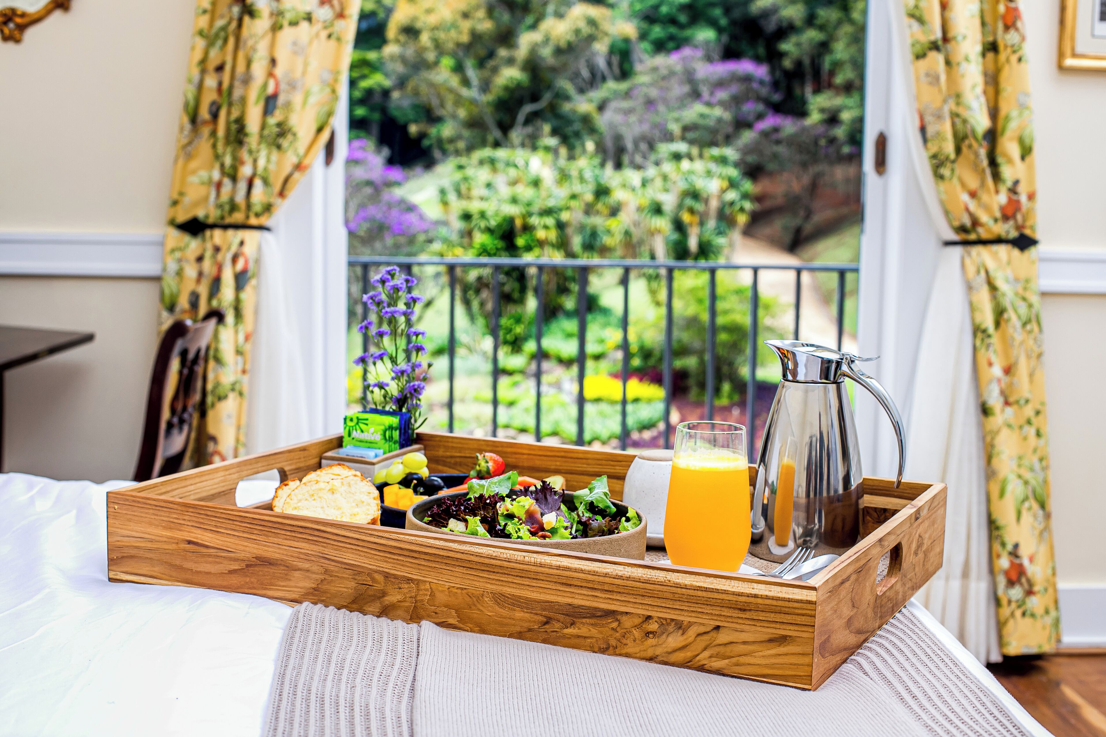 Bandeja de desayuno con ensalada, pan y jugo de naranja sobre una cama, con vistas a un jardín a través de una ventana.
