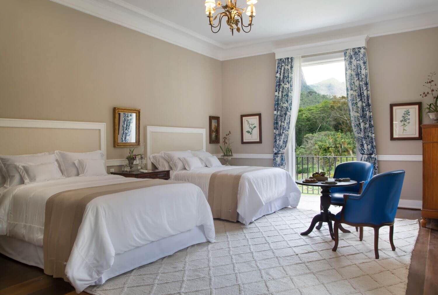 Quarto de hotel com duas camas, um lustre, varanda com cadeiras azuis e obras de arte emolduradas nas paredes.