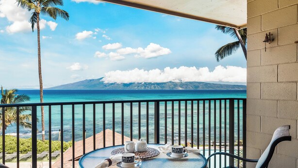 Un balcón con una mesa dispuesta para té, con vista a una playa y al océano, con palmeras y montañas a lo lejos.