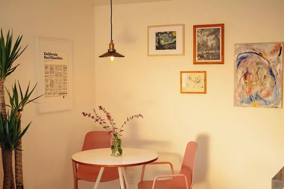 A dining area with a round table, two chairs, a hanging light fixture, and various framed artworks on the wall.