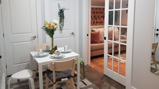 A dining area with a table set for two, a vase with flowers, and a view into a living room with a sofa and cushions. - East End Suite (Eastern Passage)