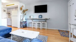 A modern living room with a white coffee table, blue sofa, and a kitchen area with white cabinets and a white refrigerator.