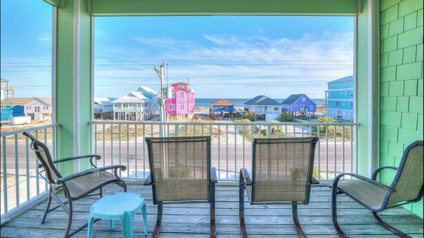 Varanda com duas cadeiras de vime e um banquinho pequeno, com vista para uma praia com casas coloridas.