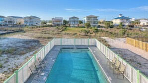 Piscina com cadeiras, cercada por uma grade, e casas à beira-mar ao fundo.