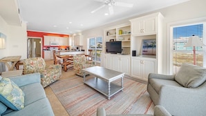 A modern living room with a dining area, kitchen, and a balcony with a view. - Holiday Summer Sale! Sundancer E/W. 10BR. Sleeps 36. Beachfront. Pool! PERFECT location. 😎 (Gulf Shores)