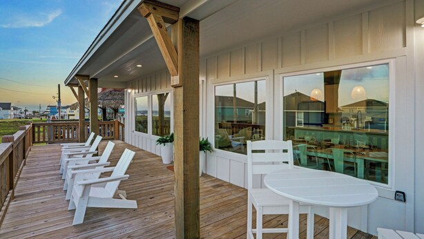 A covered outdoor area with wooden deck chairs and a table, overlooking a dining area with large windows. - Steps to Sand + Gulf Views, 2 Kings, Firepit, Dog Friendly (Crystal Beach)