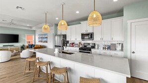 A modern kitchen with white cabinets, a large island with bar stools, and pendant lighting. - Steps to Sand + Gulf Views, 2 Kings, Firepit, Dog Friendly (Crystal Beach)