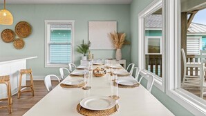 A dining room with a long table set for six, white chairs, and a view of the outside.