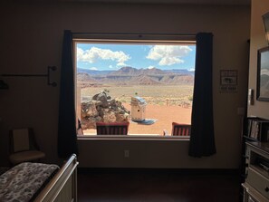 A room with a large window offering a view of a desert landscape, a small table with chairs, and a bed with a patterned bedspread.