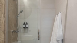 Ducha envidriada amb plat de dutxa manual i aixeta fixada a la paret. Hi ha dues ampolles de xampú i condicionador a la paret. Una tovallola blanca penja d'un ganxo a la dreta.