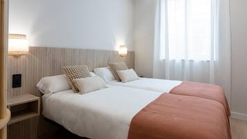 Een tweepersoonsbed met witte lakens en een bruine plaid, twee nachtkastjes met lampen, en een raam met dunne gordijnen.