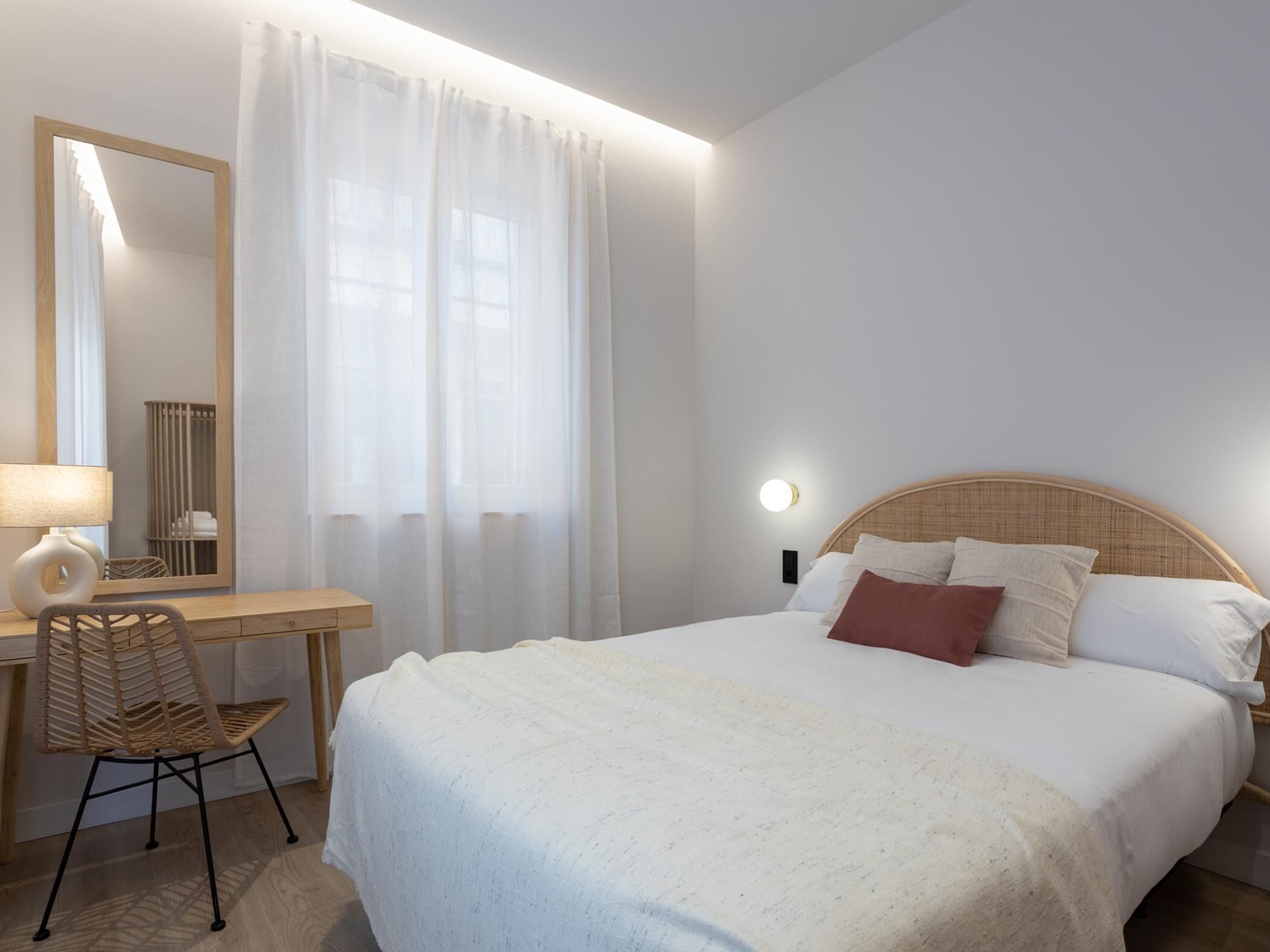 A hotel room with a bed, a desk with a chair, a mirror, and a window with curtains.