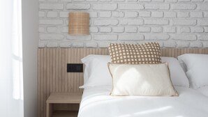A neatly made bed with white bedding and two patterned pillows, a wooden nightstand with a power outlet, and a white brick-patterned wall. - Meraki Suites (2 Rooms) (Astorga)