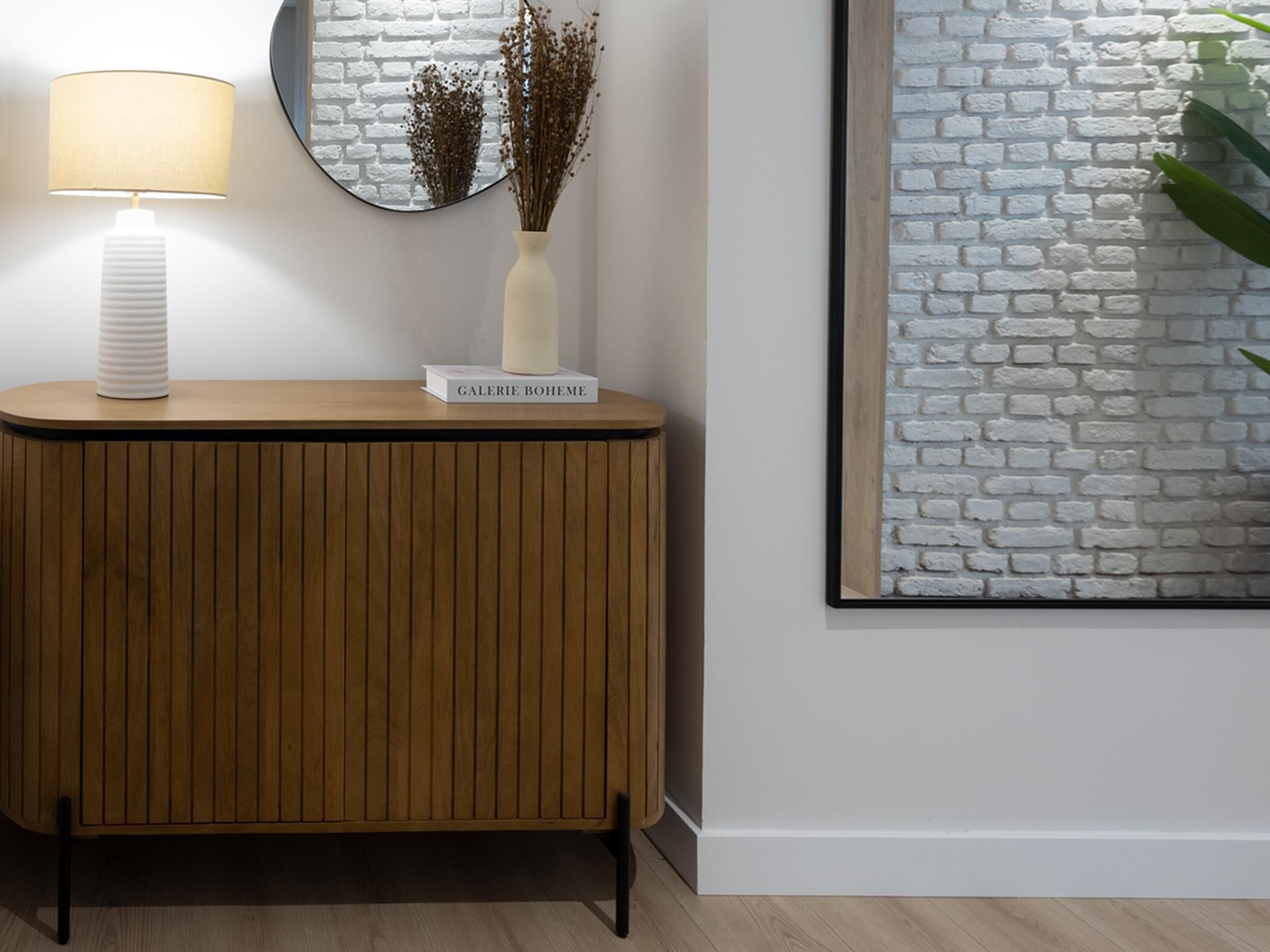 Una consola de madera con una lámpara blanca y un florero con flores secas. En la pared a la derecha, hay una obra de arte con un diseño de ladrillo blanco.