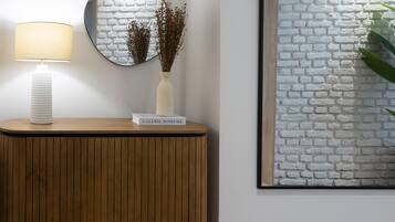 Una consola de madera con una lámpara blanca y un florero con flores secas. En la pared a la derecha, hay una obra de arte con un diseño de ladrillo blanco.