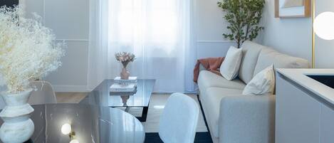 A modern living room with a glass coffee table, a sofa, and a dining table. There is a plant on the right side and a vase with flowers on the left.