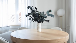 A round wooden table with a white vase containing green foliage, surrounded by grey upholstered chairs. - Meraki Suites (2 Rooms) (Astorga)