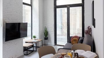 Een moderne woonruimte met een flatscreen-tv, een eethoek met tafel en stoelen, een bank en een groot raam met uitzicht op de straat.