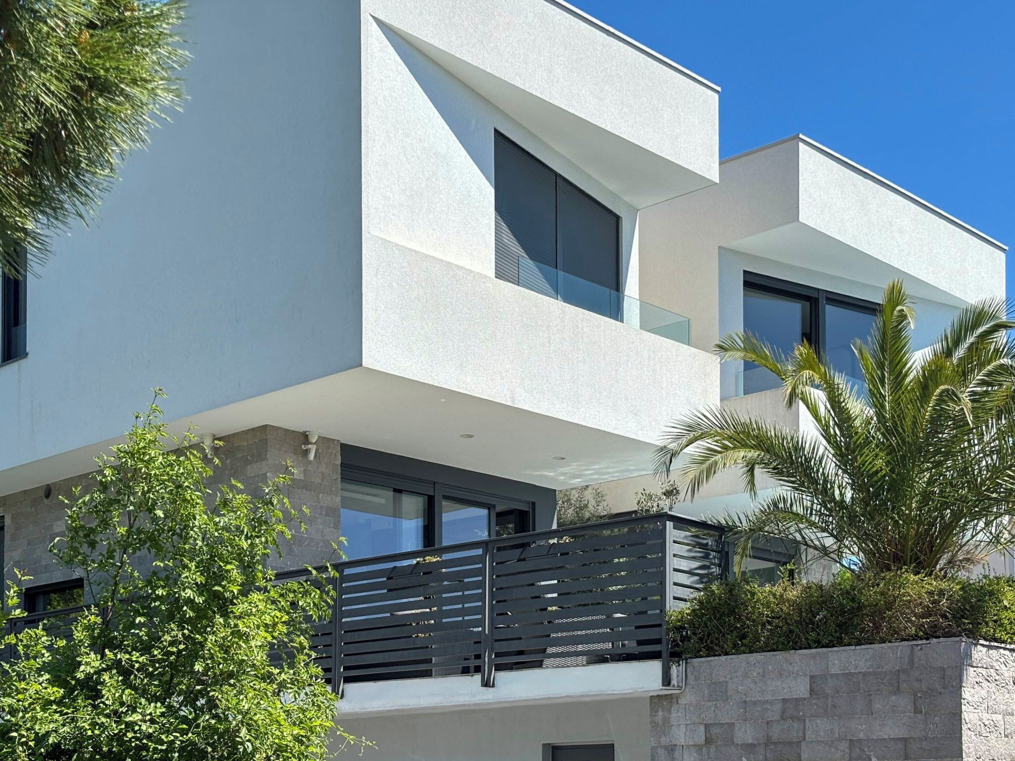 A modern two-story building with large windows, a balcony, and surrounding greenery.