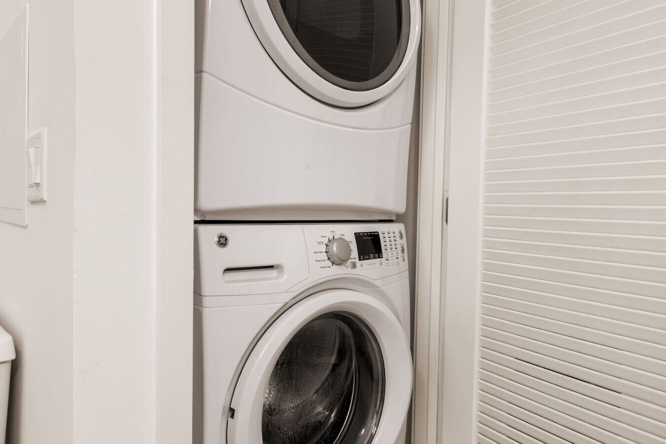 A stacked washer and dryer unit in a laundry room with a white exterior and a closed shutter door.