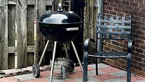 Una parrilla negra con un soporte metálico, ubicada junto a una silla de metal en un patio trasero con cerca de madera y pared de ladrillo.